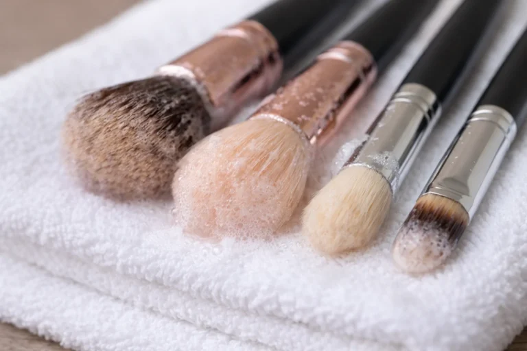 Pinceaux de maquillage propres en cours de nettoyage posés sur une serviette, avec mousse et eau, illustrant l’entretien des pinceaux maquillage.